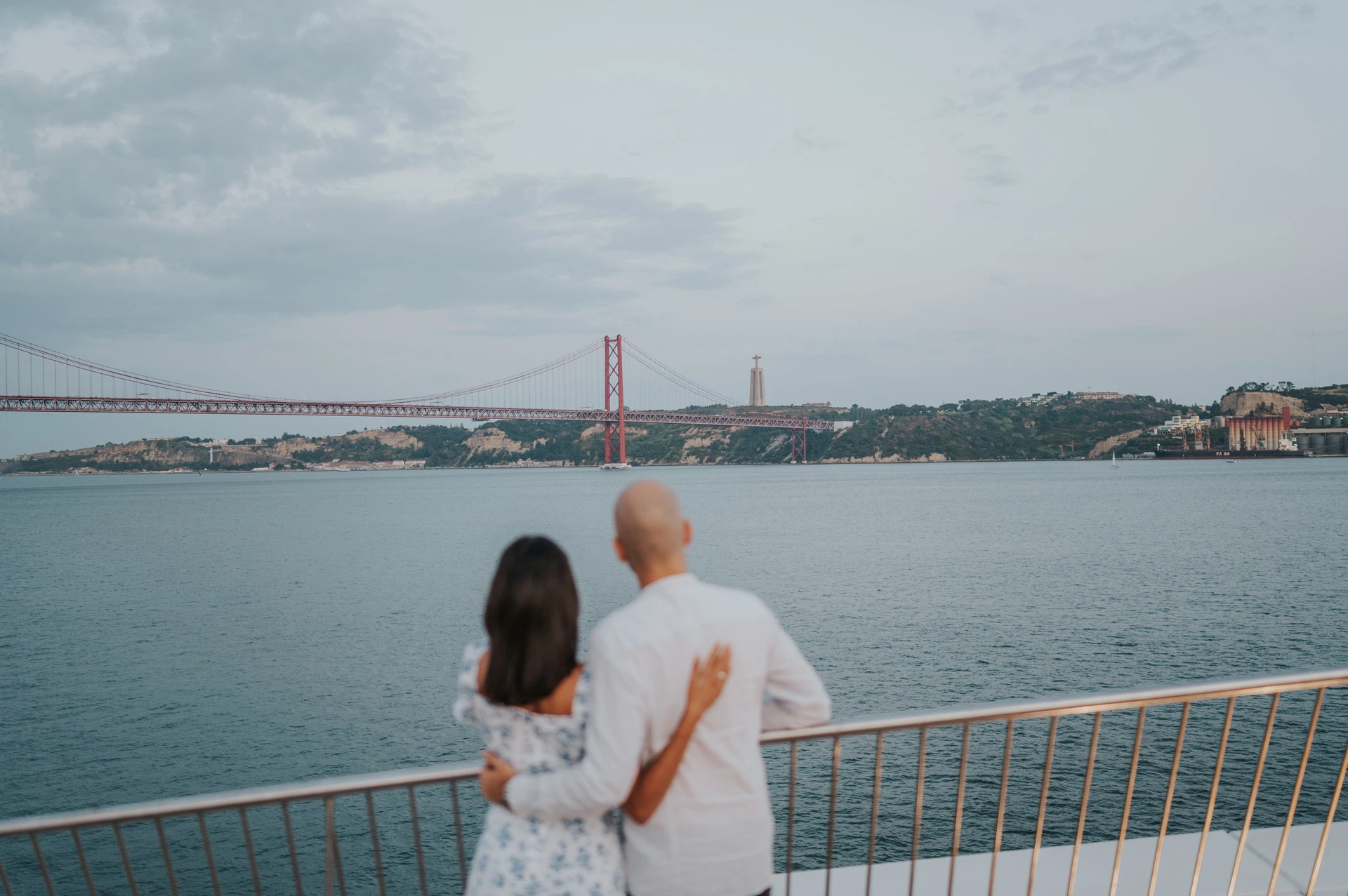 Looking out over the river together | Lisbon couple photoshoot The couple seen from behind with their arms around each other, looking out over the river and 25 de Abril Bridge during their couple photoshoot in Lisbon.
