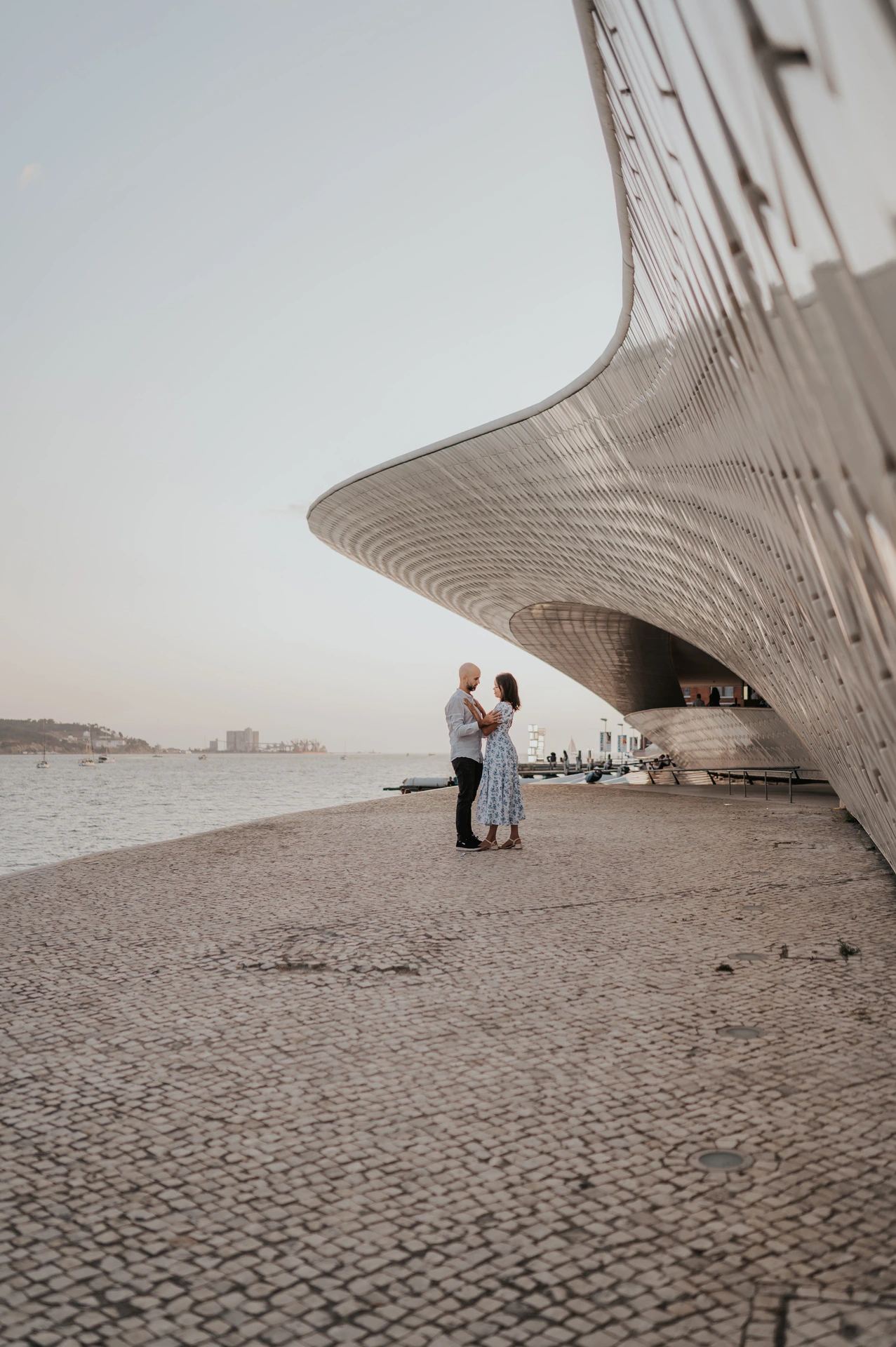 A distant portrait of the couple embracing beneath the curved MAAT facade during their couple photoshoot in Lisbon.