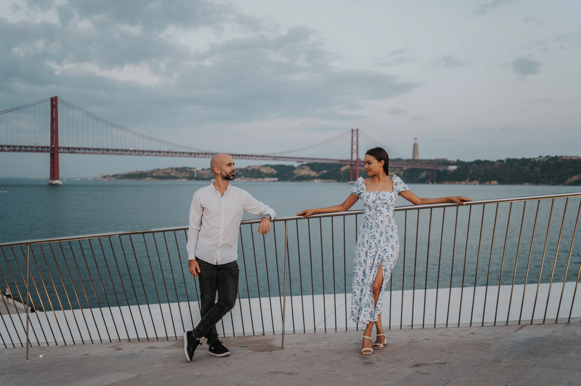 A wide portrait of the couple leaning against the railing with the river and bridge behind them during their couple photoshoot in Lisbon.