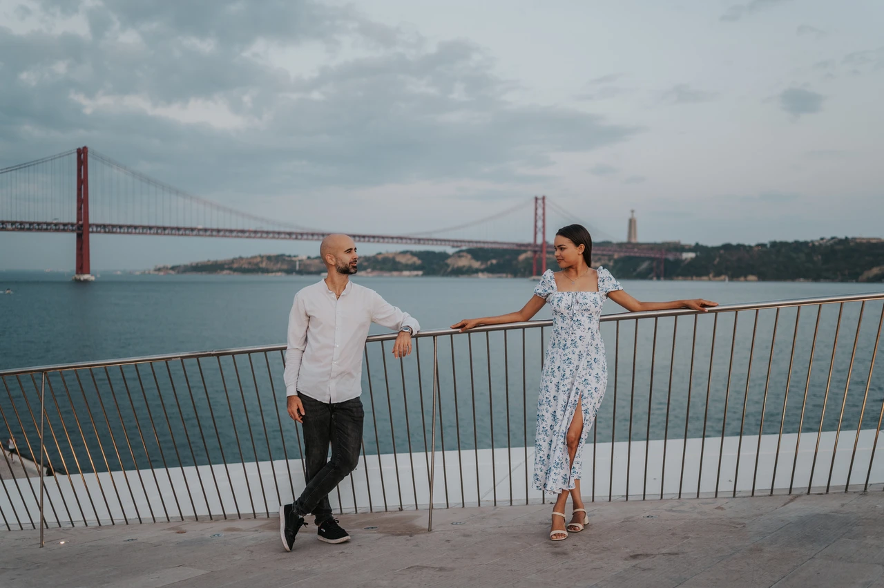 Together at the river railing | Lisbon couple photoshoot A wide portrait of the couple leaning against the railing with the river and bridge behind them during their couple photoshoot in Lisbon.