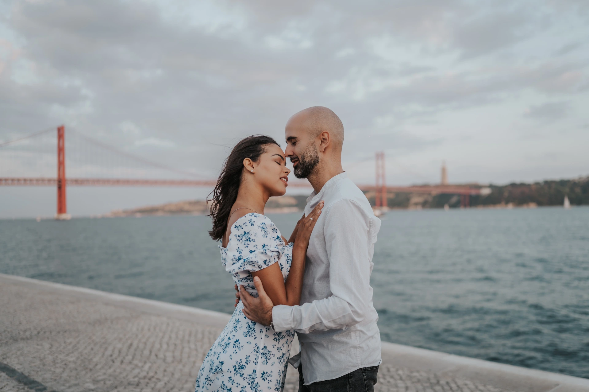 The couple holding each other close with the river and 25 de Abril Bridge behind them during their couple photoshoot in Lisbon.