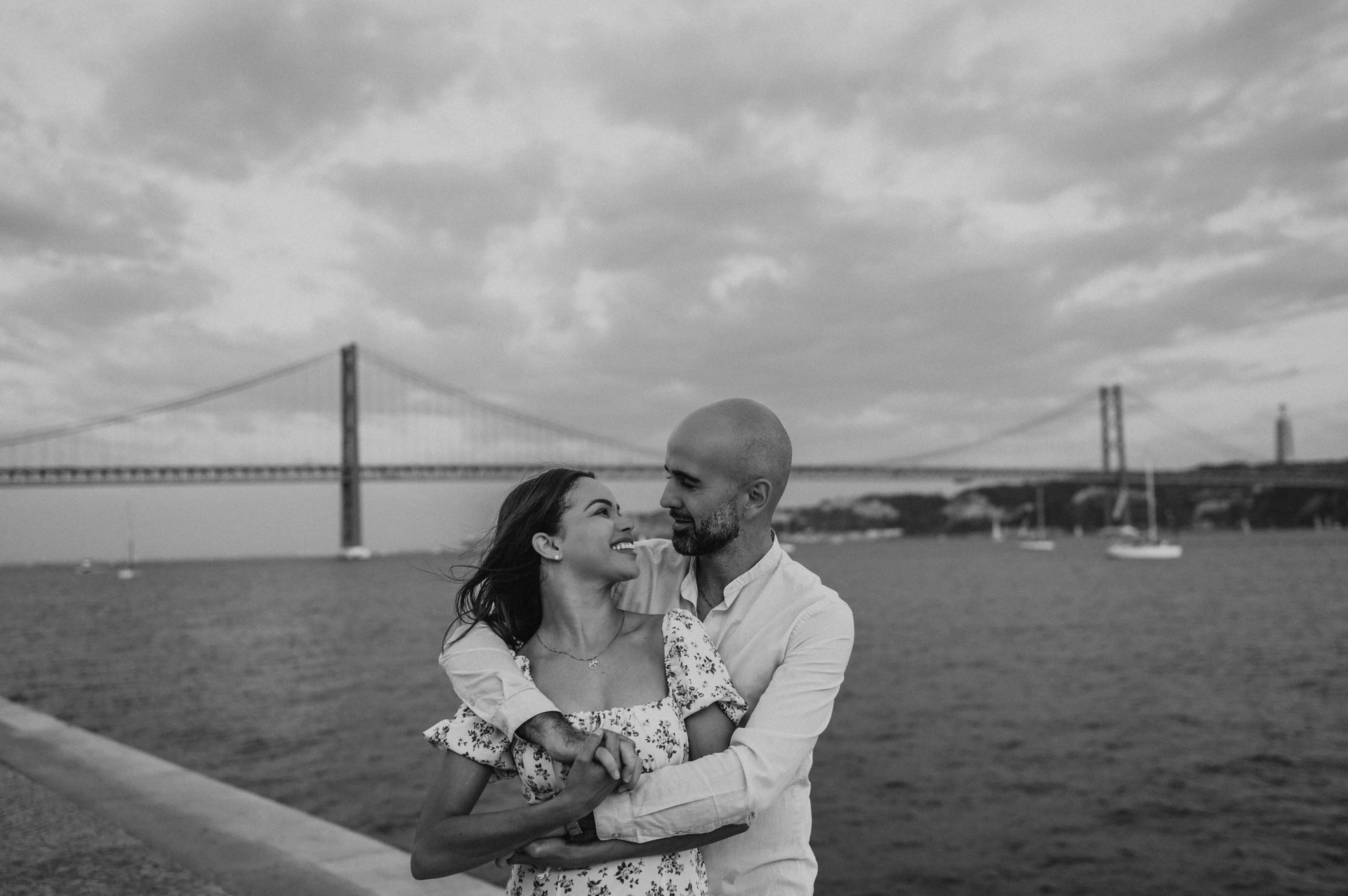 A black-and-white photograph of the couple smiling in an embrace beside the river during their couple photoshoot in Lisbon.