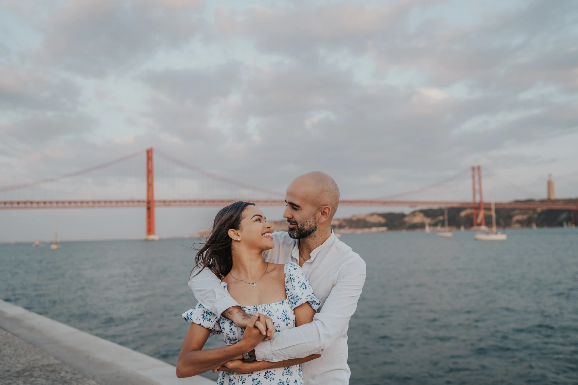 The woman smiling up at her partner while he holds her beside the river during their couple photoshoot in Lisbon.