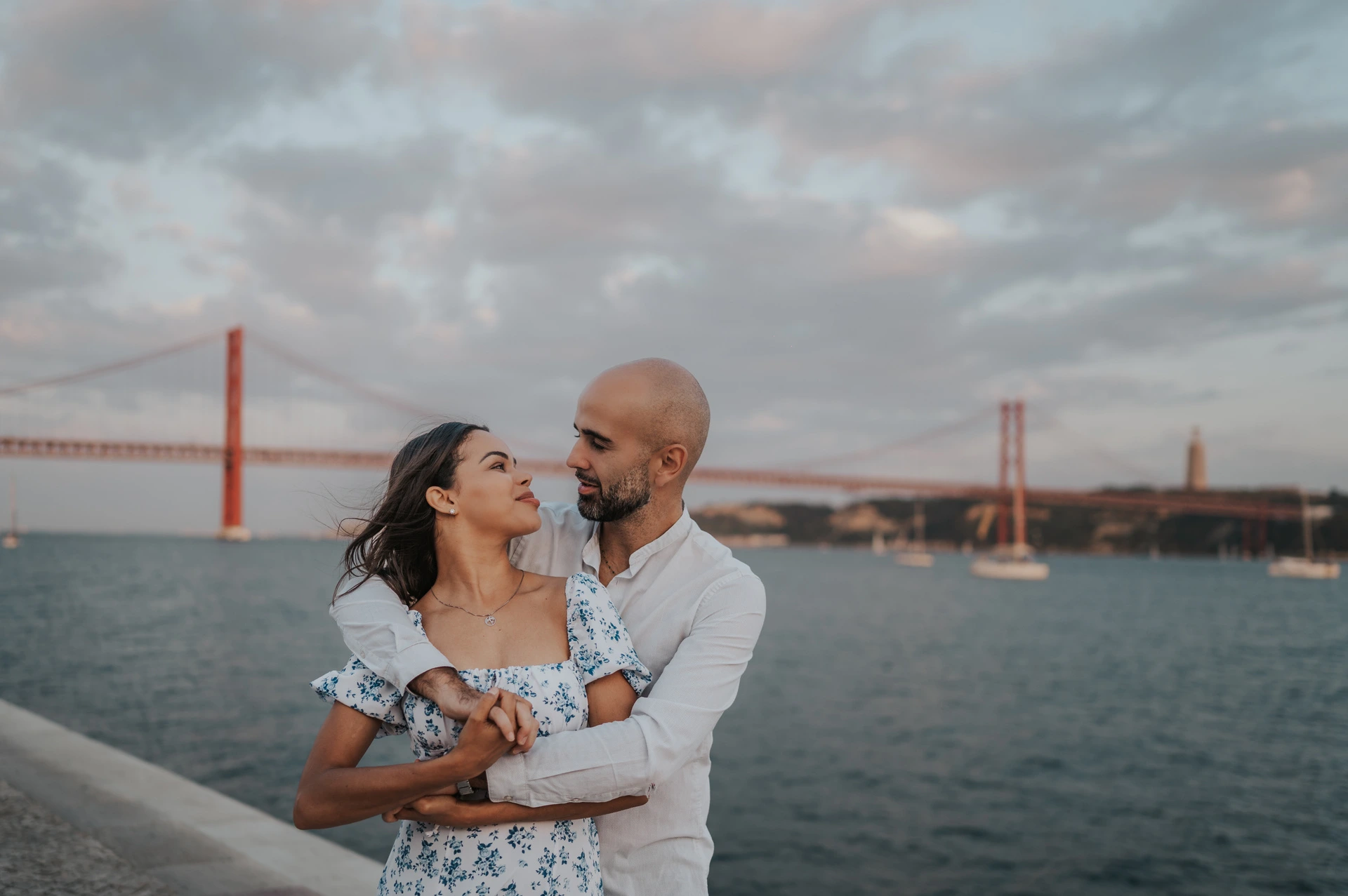 An embrace with the bridge behind them | Lisbon couple photoshoot The couple looking at one another as he embraces her from behind beside the river during their couple photoshoot in Lisbon.