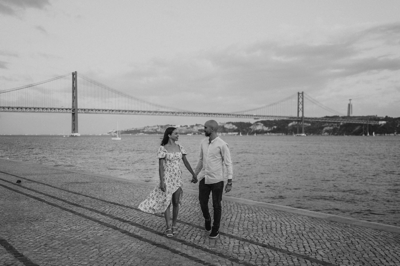 A waterfront walk in black and white | Lisbon couple photoshoot A black-and-white photograph of the couple holding hands beside the river and 25 de Abril Bridge during their couple photoshoot in Lisbon.