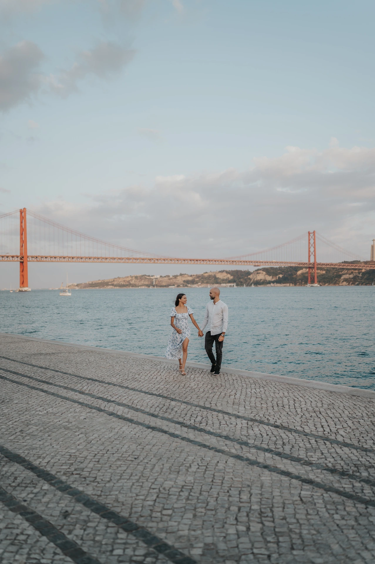 A distant view of the couple walking hand in hand beside the river with the 25 de Abril Bridge behind them during their couple photoshoot in Lisbon.