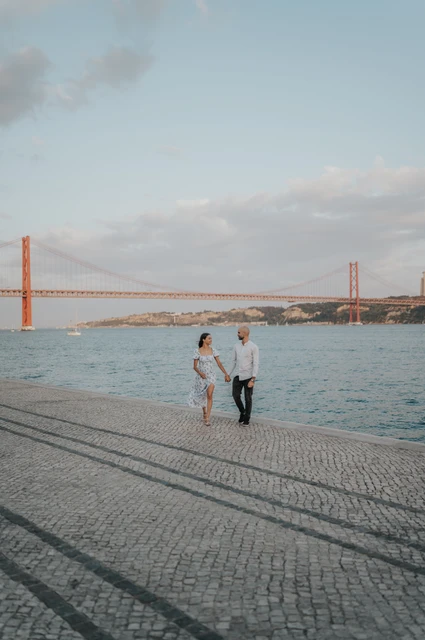 A river walk beneath the evening sky | Lisbon couple photoshoot A distant view of the couple walking hand in hand beside the river with the 25 de Abril Bridge behind them during their couple photoshoot in Lisbon.