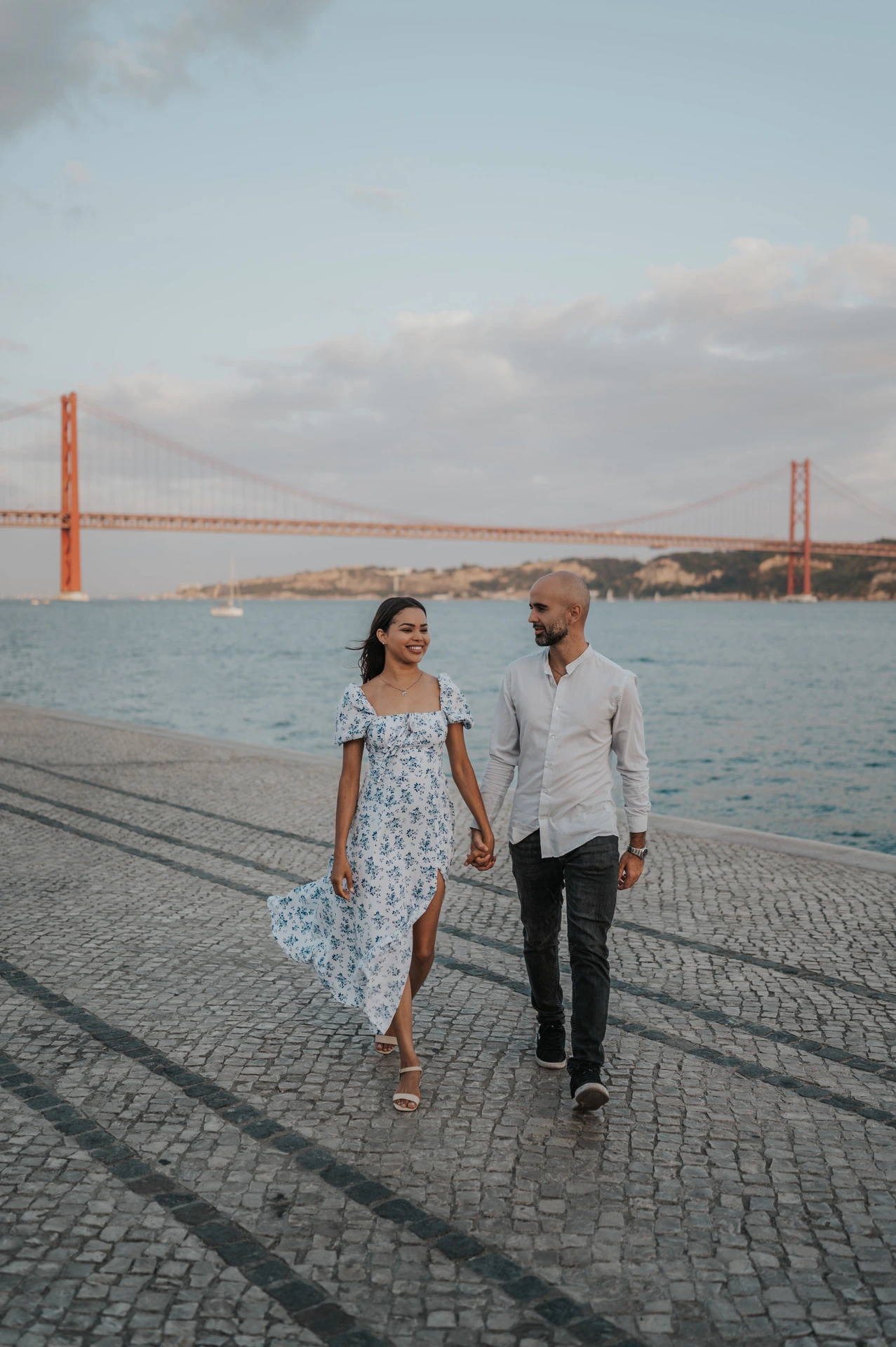 The woman’s floral dress moving as the couple walk along the cobbled waterfront during their couple photoshoot in Lisbon.