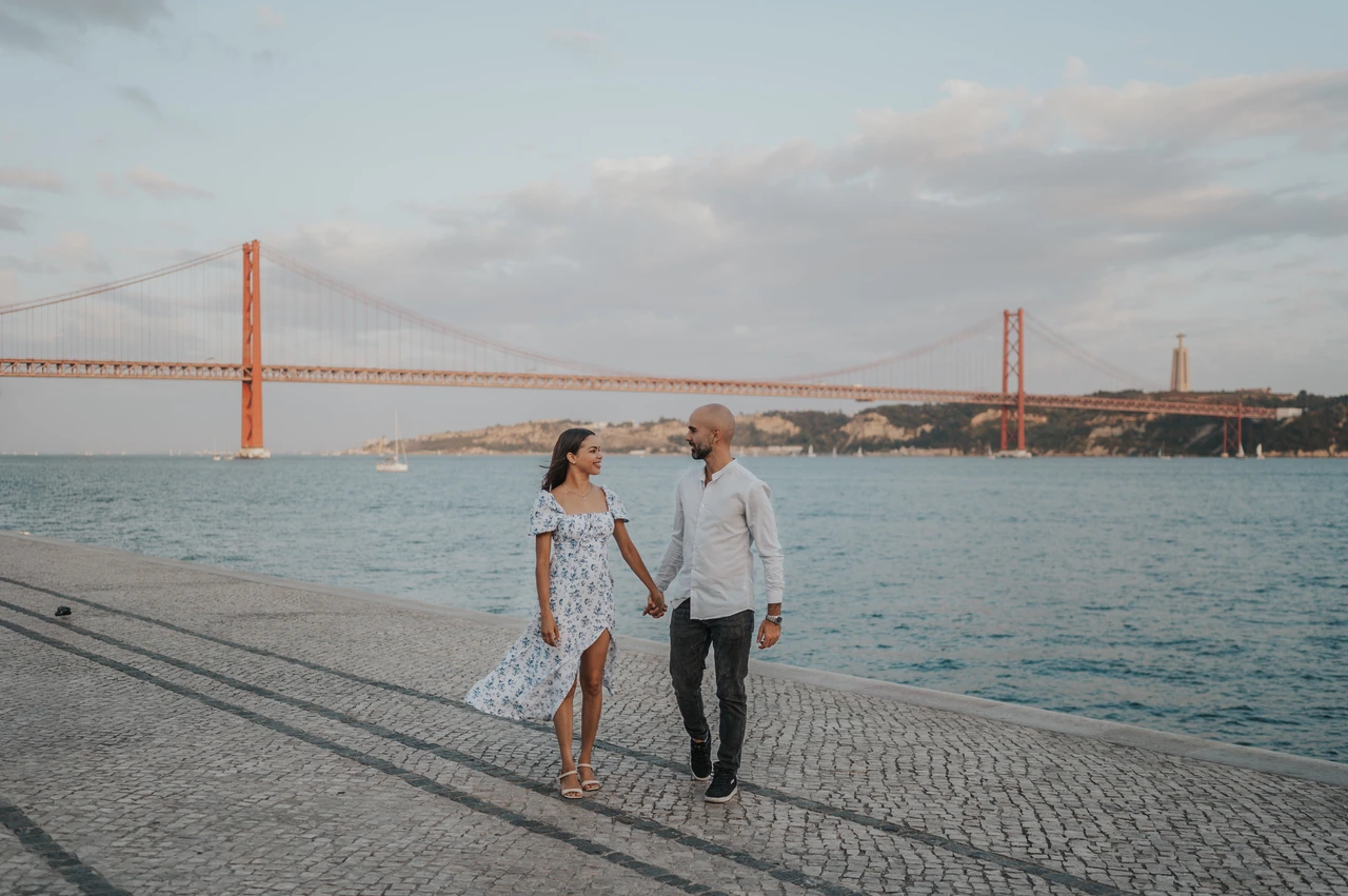 Hand in hand along the river | Lisbon couple photoshoot A wide photograph of the couple holding hands beside the river and 25 de Abril Bridge during their couple photoshoot in Lisbon.