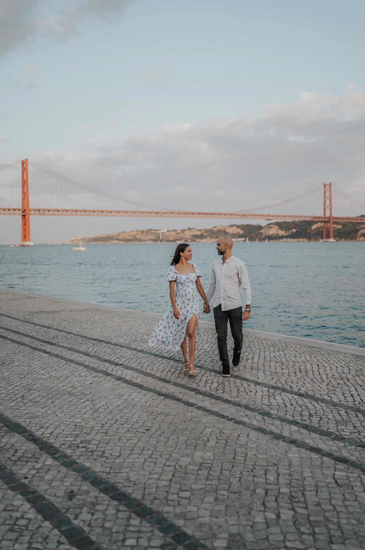 The couple walking towards the camera with the river and 25 de Abril Bridge behind them during their couple photoshoot in Lisbon.