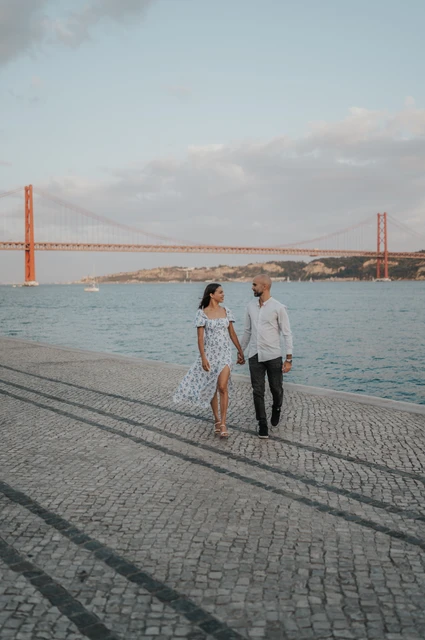 Walking beside the 25 de Abril Bridge | Lisbon couple photoshoot The couple walking towards the camera with the river and 25 de Abril Bridge behind them during their couple photoshoot in Lisbon.