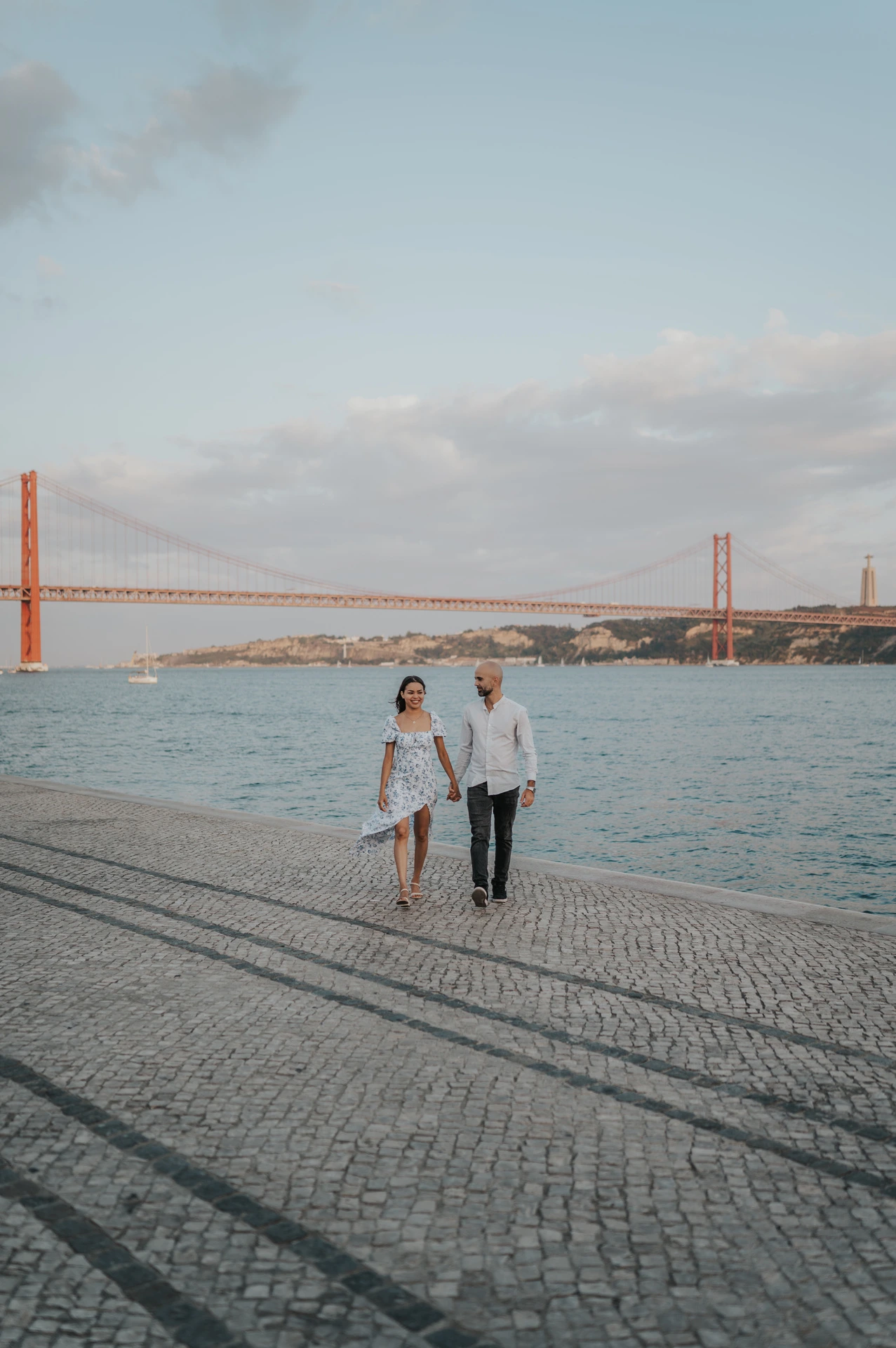 The couple walking together beside the water beneath a wide sky and the 25 de Abril Bridge during their couple photoshoot in Lisbon.