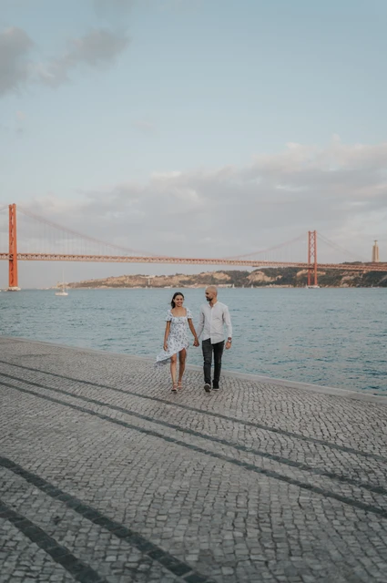 Wide skies over a waterfront walk | Lisbon couple photoshoot The couple walking together beside the water beneath a wide sky and the 25 de Abril Bridge during their couple photoshoot in Lisbon.
