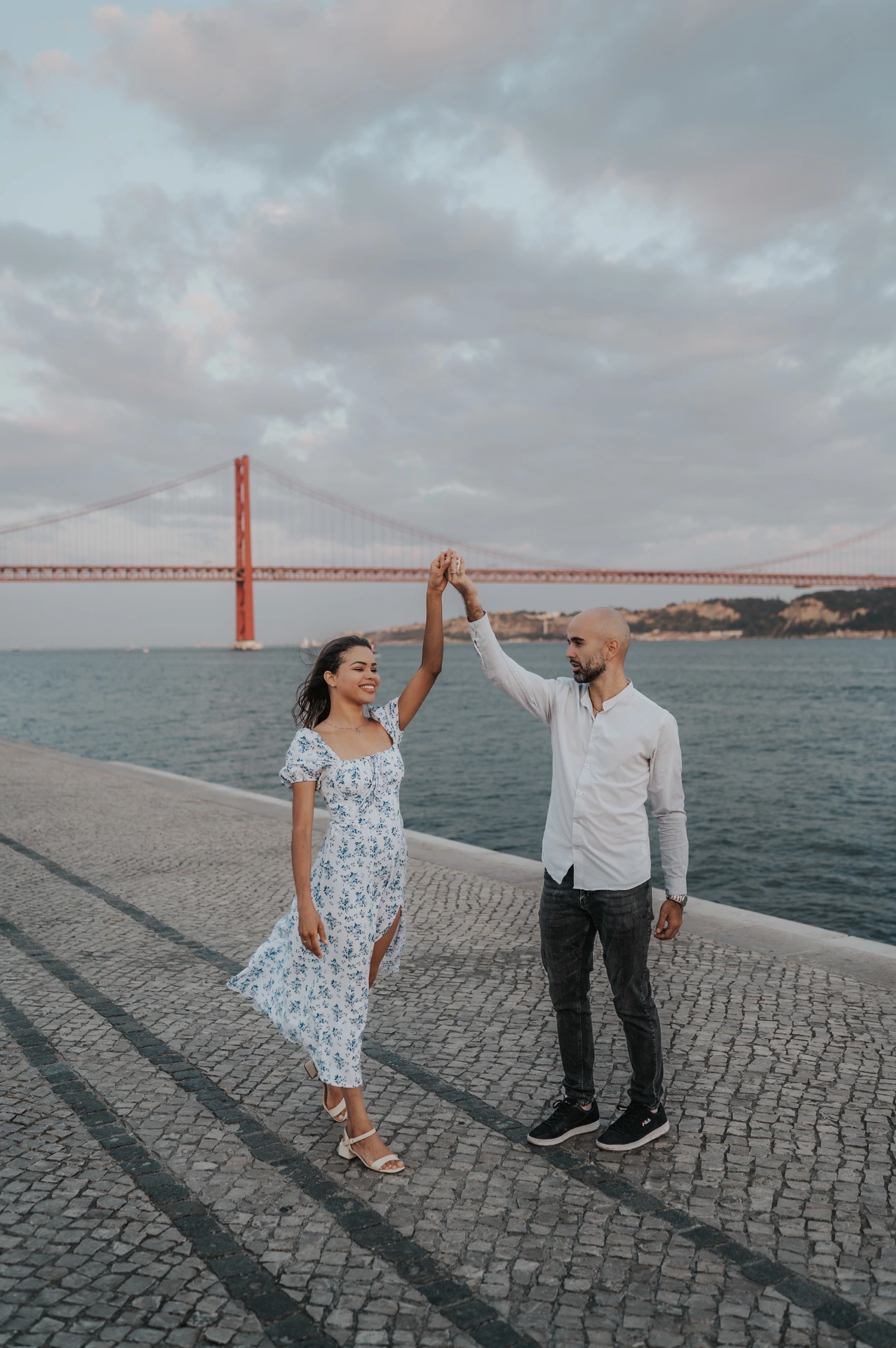 The couple raising their joined hands for a turn beside the river and bridge during their couple photoshoot in Lisbon.