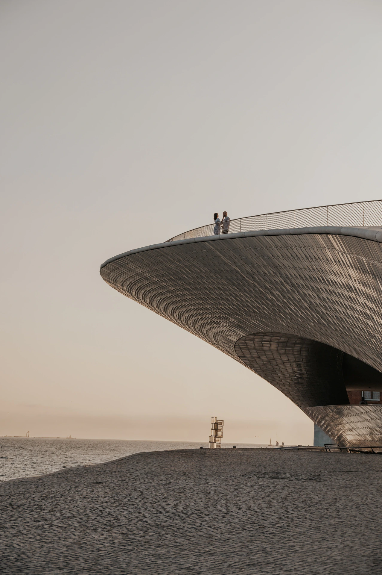 The couple standing together above the curved MAAT facade with open sky behind them during their couple photoshoot in Lisbon.