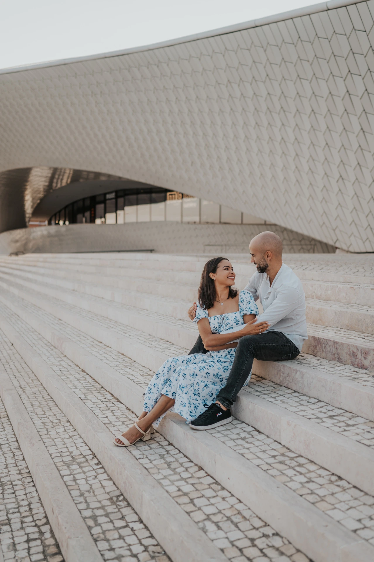 The woman smiling up at her partner as they sit together on the MAAT steps during their couple photoshoot in Lisbon.