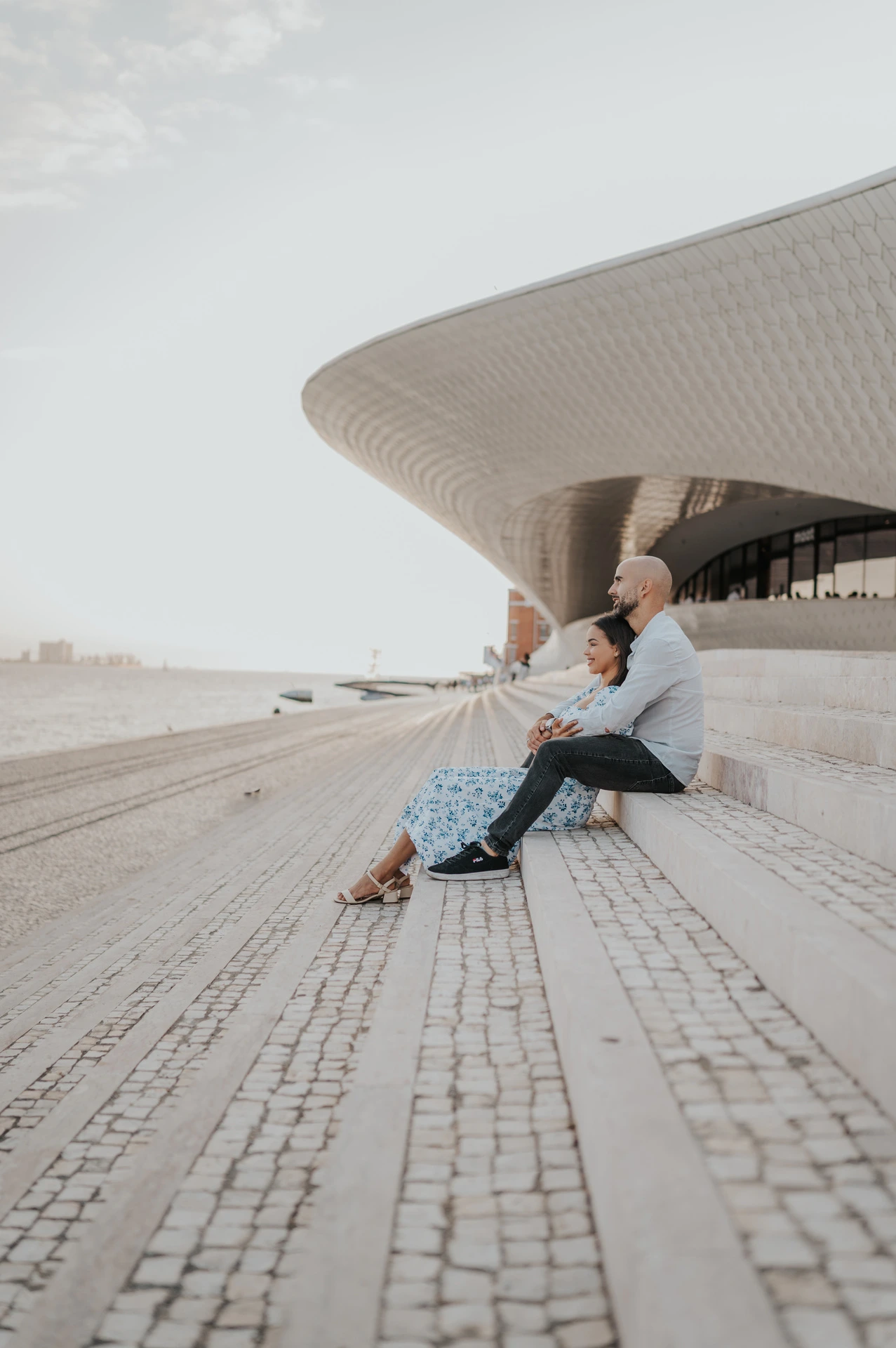 A side view of the couple seated on the MAAT steps, with cobblestones leading towards them during their couple photoshoot in Lisbon.