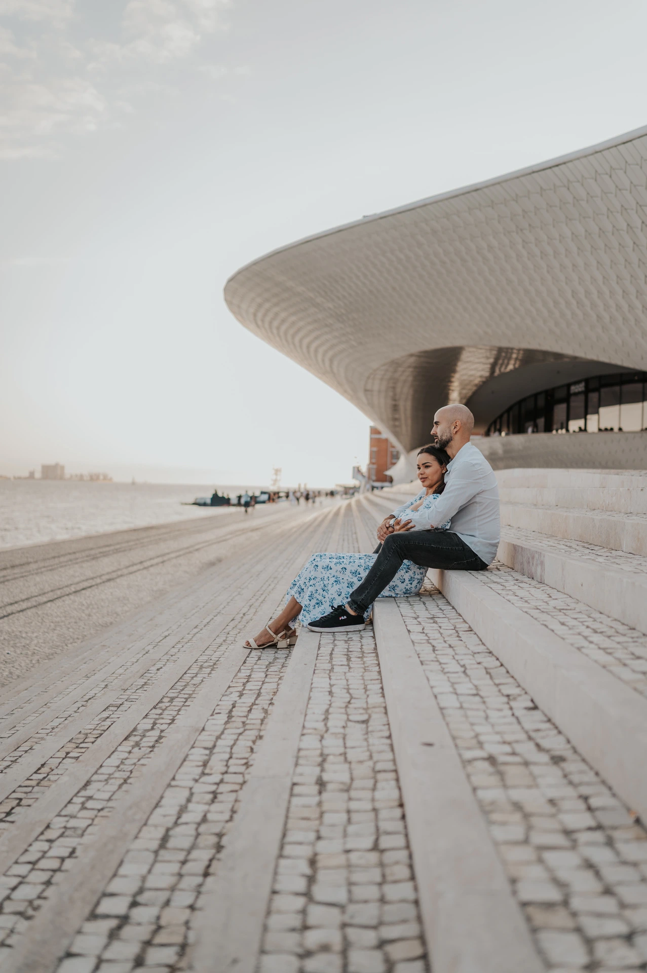 The couple sitting close together on the steps beneath the sweeping MAAT facade during their couple photoshoot in Lisbon.