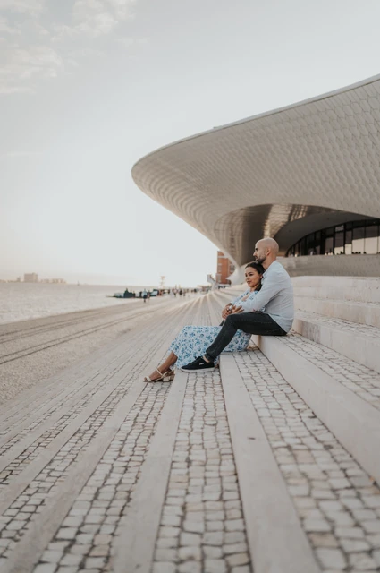 Seated beneath the sweep of the MAAT | Lisbon couple photoshoot The couple sitting close together on the steps beneath the sweeping MAAT facade during their couple photoshoot in Lisbon.