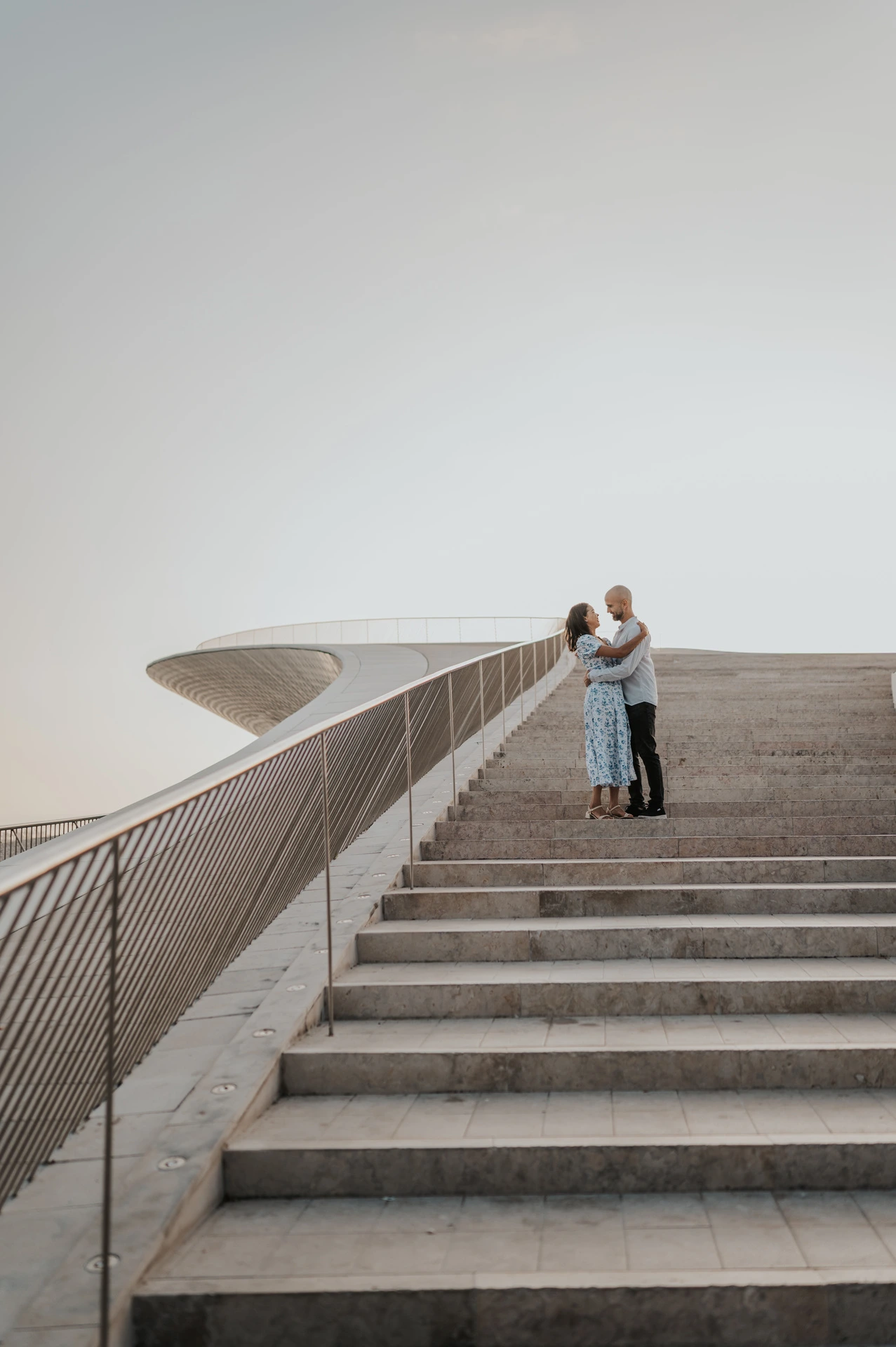 The couple embracing at the top of the MAAT staircase beside the curved railing during their couple photoshoot in Lisbon.