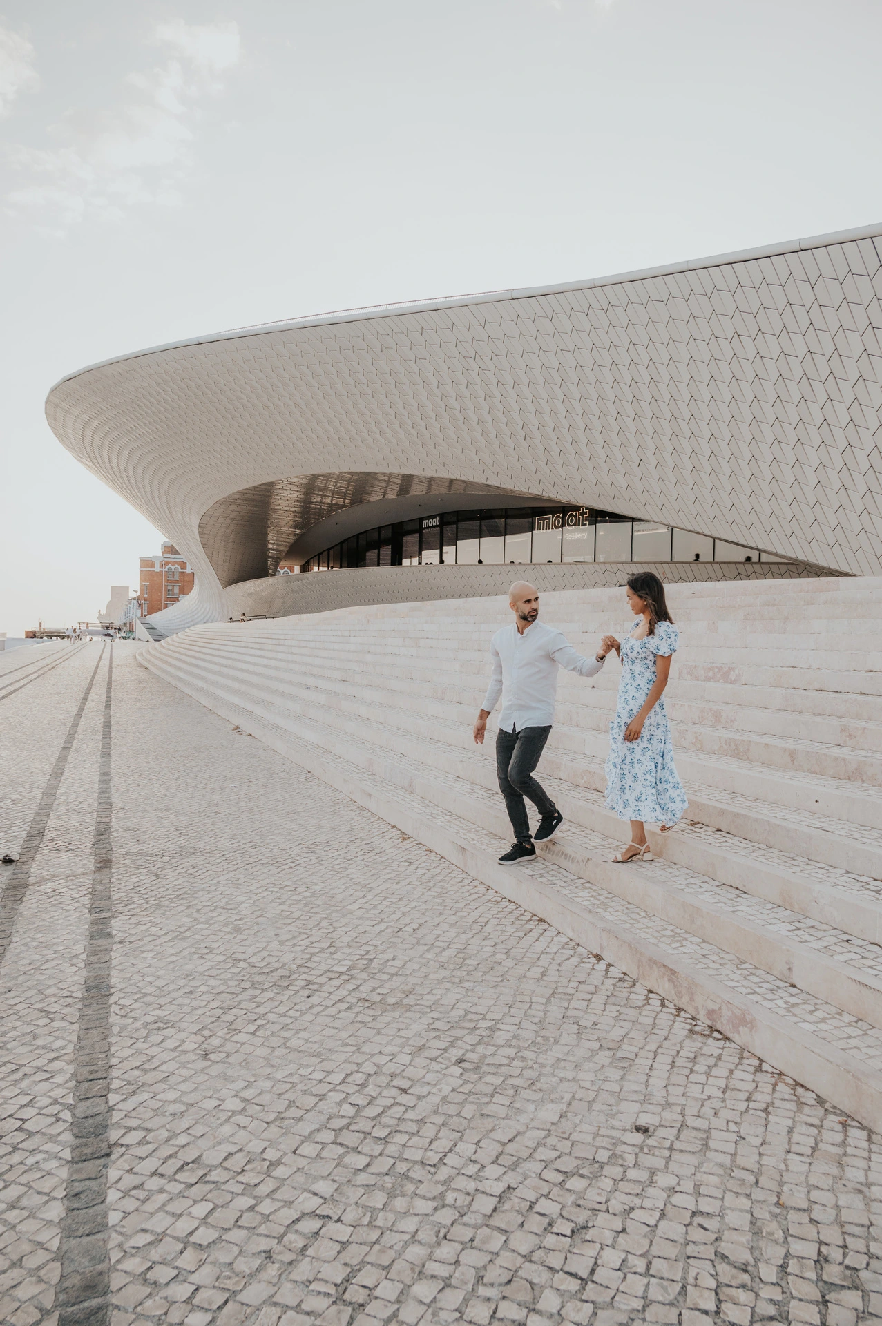 The couple standing hand in hand on the steps beneath the curved MAAT facade during their couple photoshoot in Lisbon.