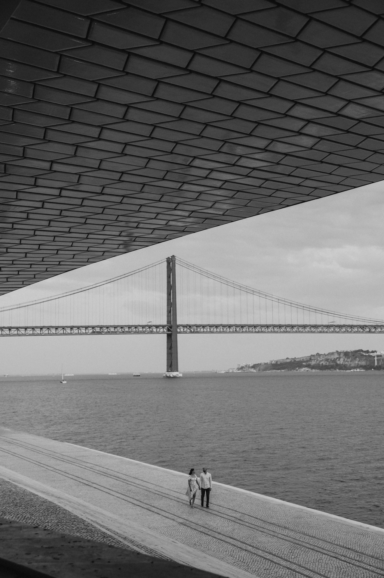 A black-and-white view of the couple beneath the MAAT roof with the bridge beyond during their couple photoshoot in Lisbon.