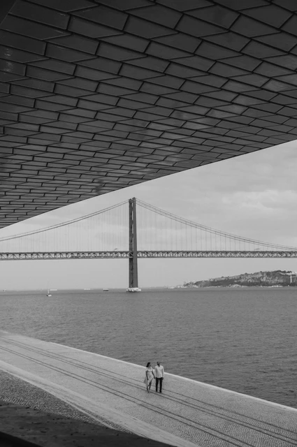 A framed bridge view in black and white | Lisbon couple photoshoot A black-and-white view of the couple beneath the MAAT roof with the bridge beyond during their couple photoshoot in Lisbon.