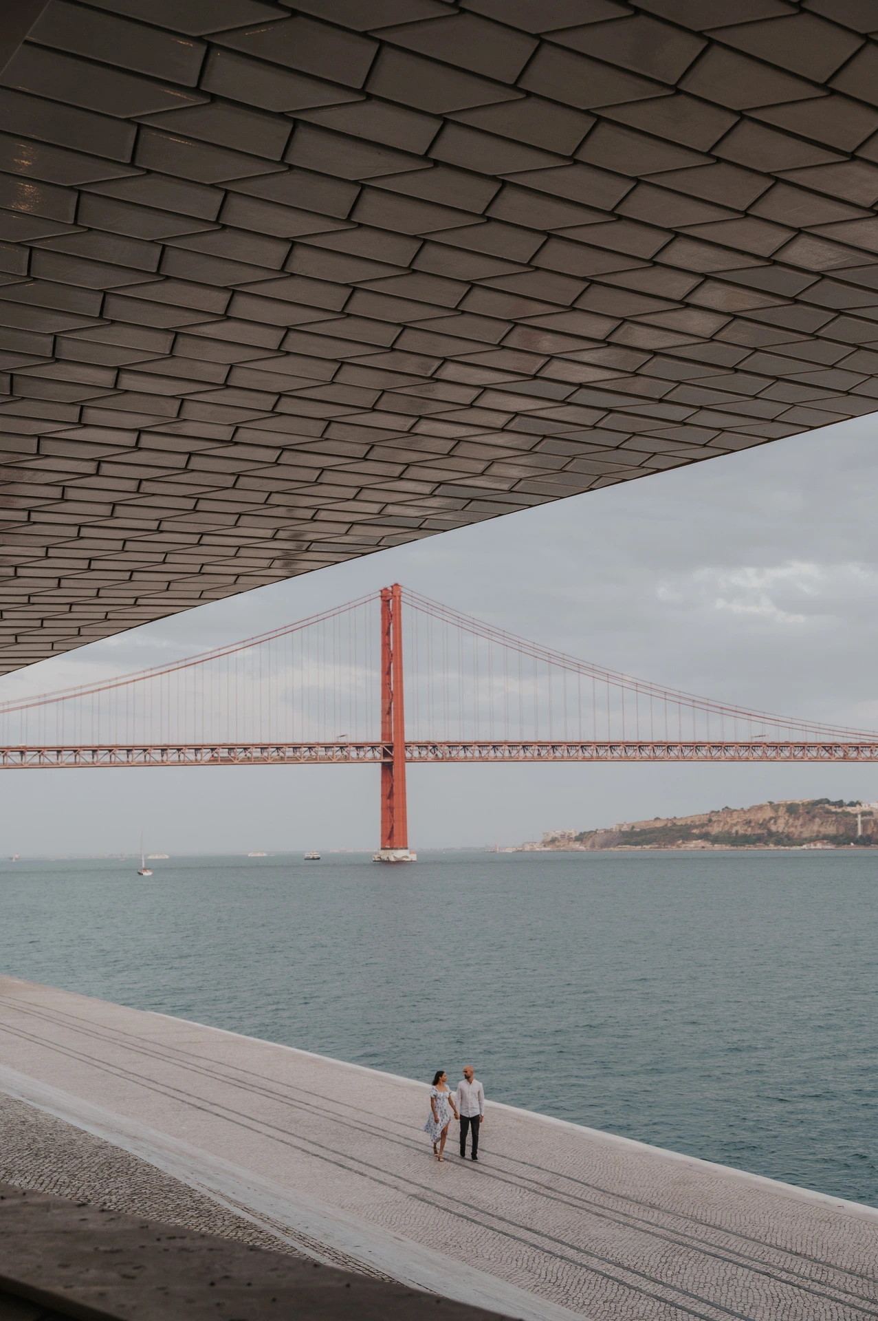 The couple standing on the waterfront beneath the MAAT roof, with the 25 de Abril Bridge framed beyond during their couple photoshoot in Lisbon.