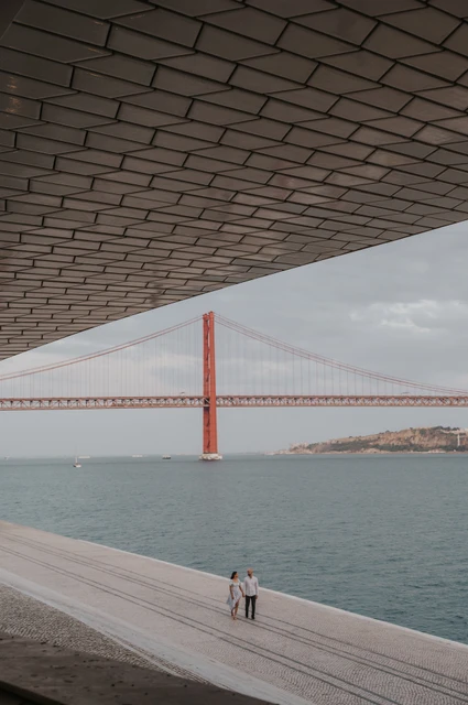 The bridge framed beneath the MAAT roof | Lisbon couple photoshoot The couple standing on the waterfront beneath the MAAT roof, with the 25 de Abril Bridge framed beyond during their couple photoshoot in Lisbon.
