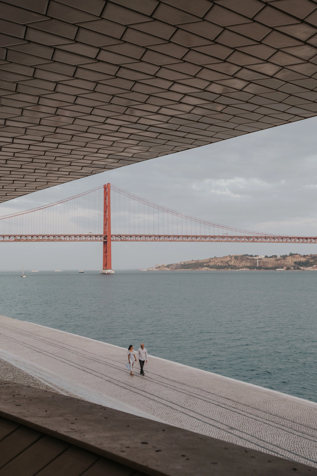 A wide view beneath the MAAT roof towards the couple on the waterfront and the 25 de Abril Bridge during their couple photoshoot in Lisbon.