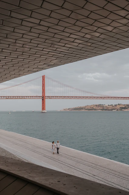 A wide river view beneath the MAAT | Lisbon couple photoshoot A wide view beneath the MAAT roof towards the couple on the waterfront and the 25 de Abril Bridge during their couple photoshoot in Lisbon.