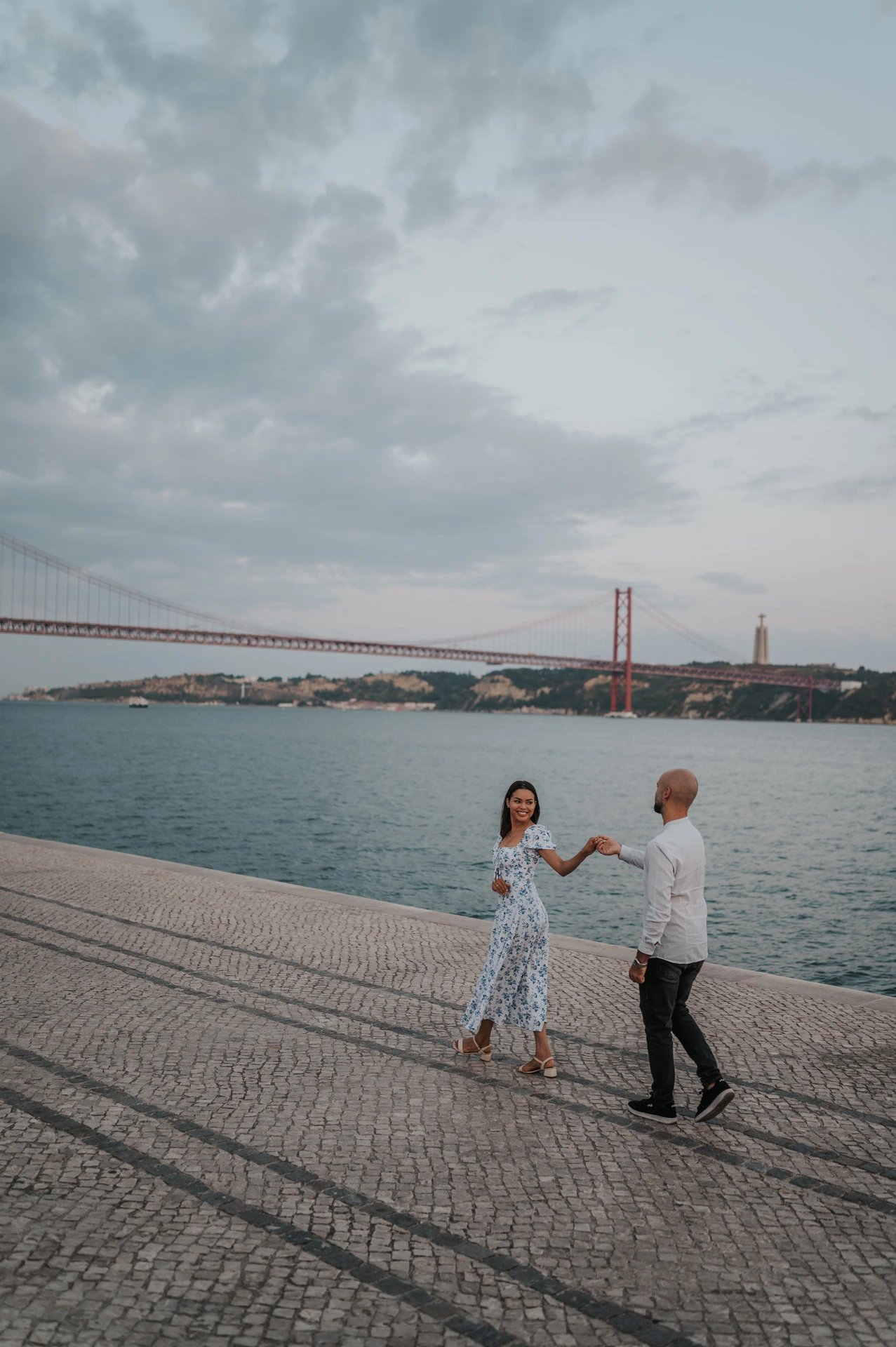 The couple holding hands with the river and bridge stretching out behind them during their couple photoshoot in Lisbon.