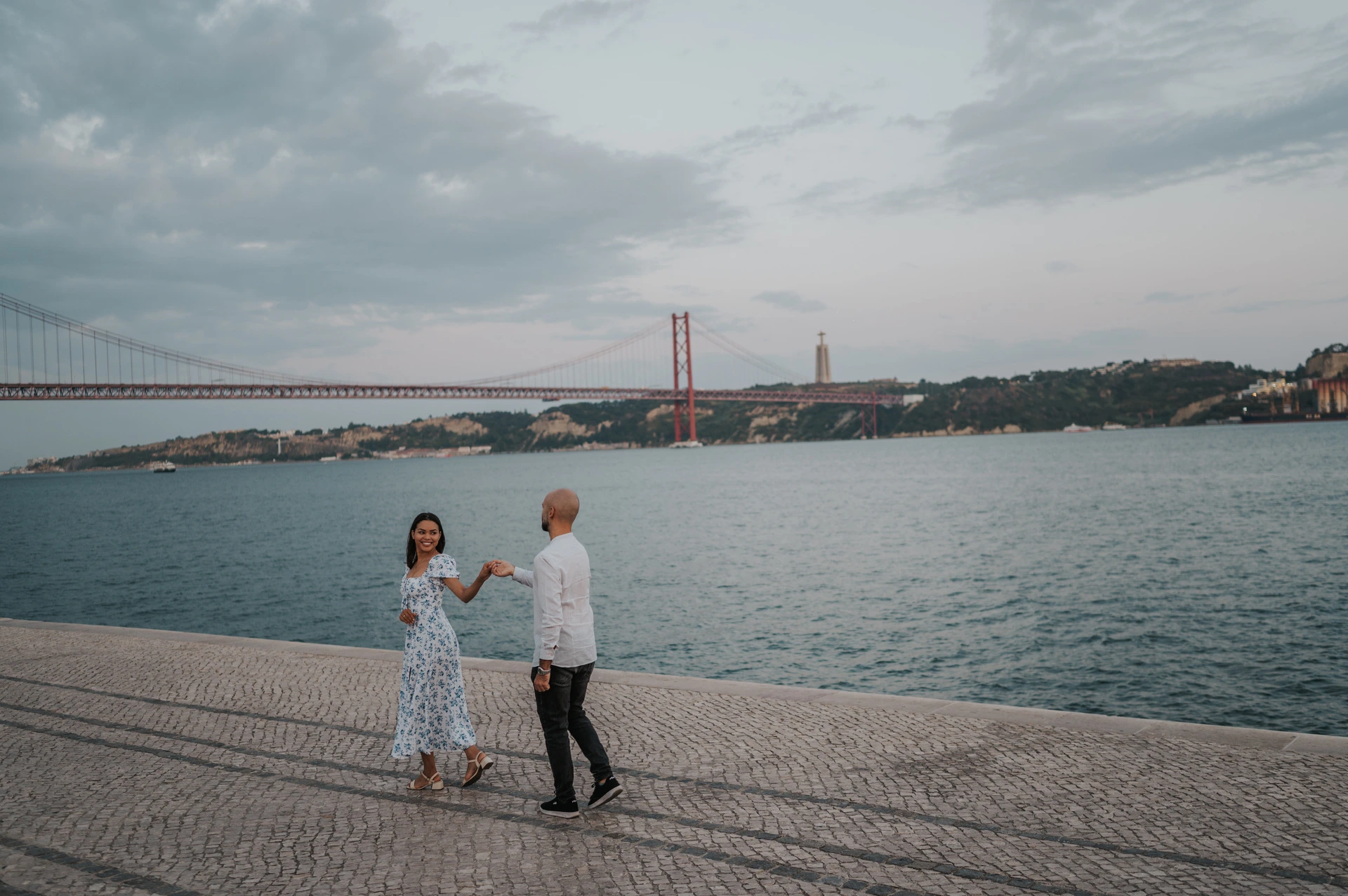 A wide portrait of the couple holding hands on the cobbled waterfront under the evening sky during their couple photoshoot in Lisbon.