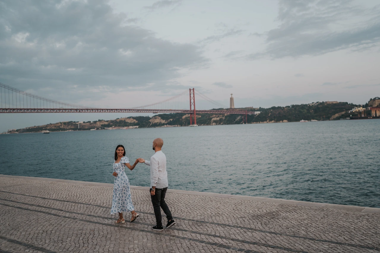 A wide portrait on the waterfront | Lisbon couple photoshoot A wide portrait of the couple holding hands on the cobbled waterfront under the evening sky during their couple photoshoot in Lisbon.
