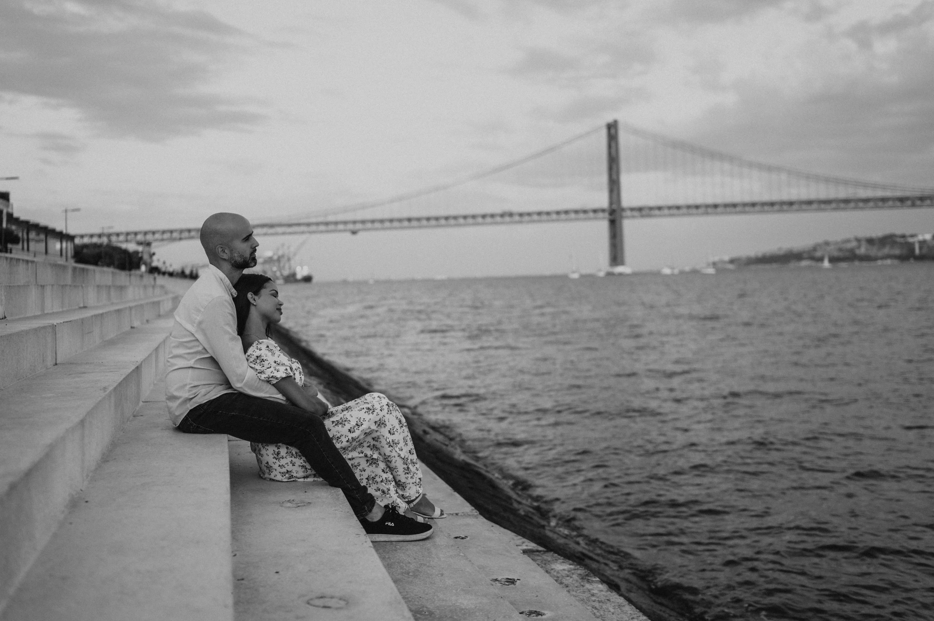 A black-and-white portrait of the couple sitting beside the river with the 25 de Abril Bridge behind them during their couple photoshoot in Lisbon.