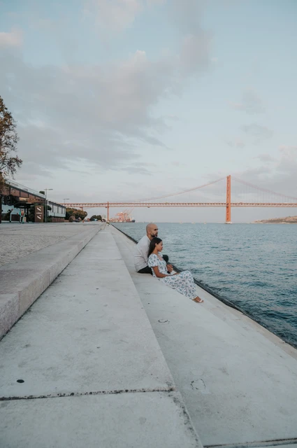 A quiet seat beside the river | Lisbon couple photoshoot The couple sitting on the broad waterfront steps with the 25 de Abril Bridge in the distance during their couple photoshoot in Lisbon.