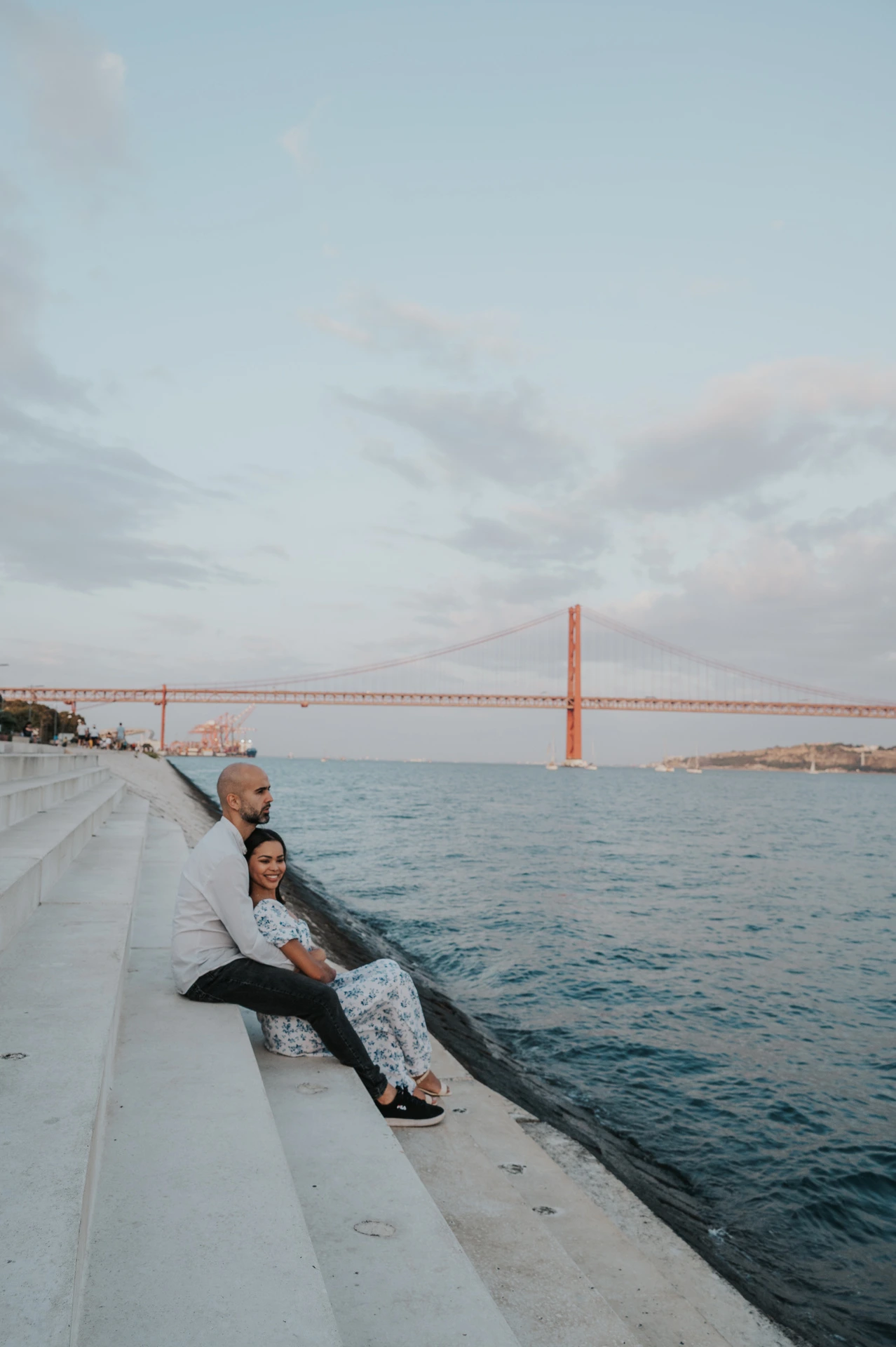 The seated couple looking towards the camera with the river and bridge behind them during their couple photoshoot in Lisbon.