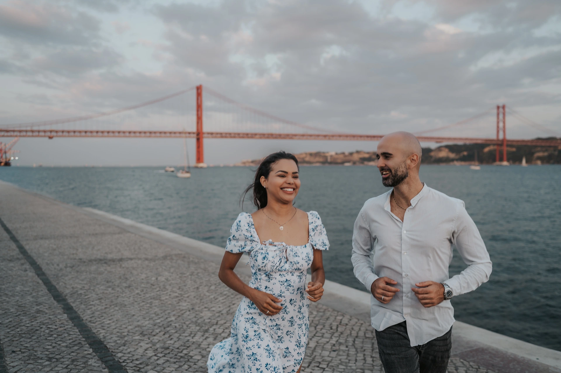 The couple smiling as they walk towards the camera beside the river during their couple photoshoot in Lisbon.