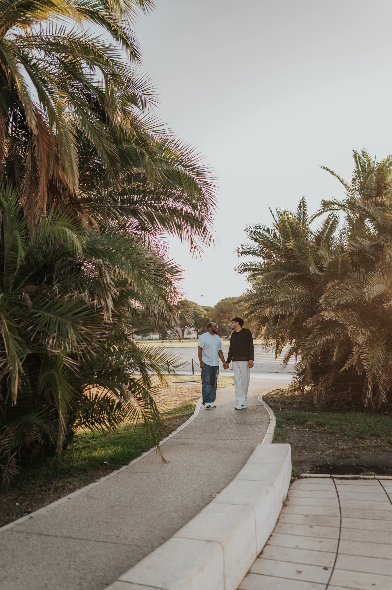 The couple walking hand in hand along a curved path between palm trees during their couple photoshoot in Lisbon.