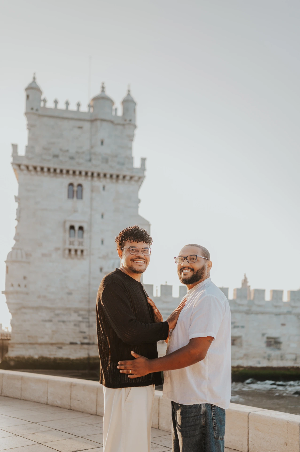 The couple holding each other and smiling towards the camera with Belém Tower behind them during their couple photoshoot in Lisbon.
