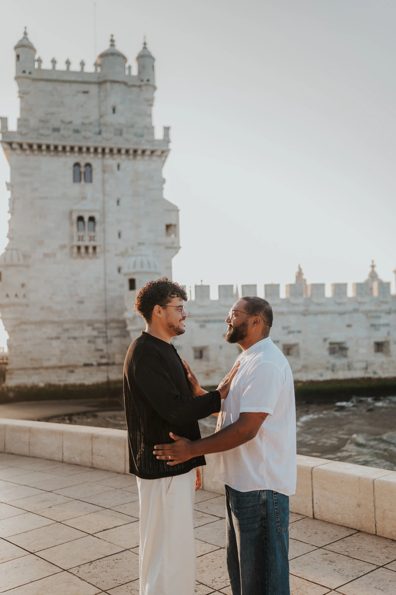 The couple facing each other and smiling with their hands resting on one another in front of Belém Tower during their couple photoshoot in Lisbon.