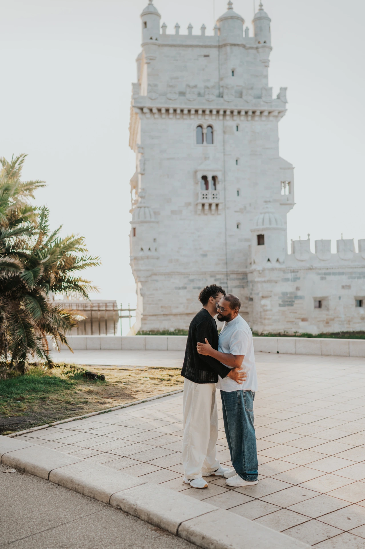 The couple embracing with Belém Tower behind them and palm leaves at the side of the frame during their couple photoshoot in Lisbon.