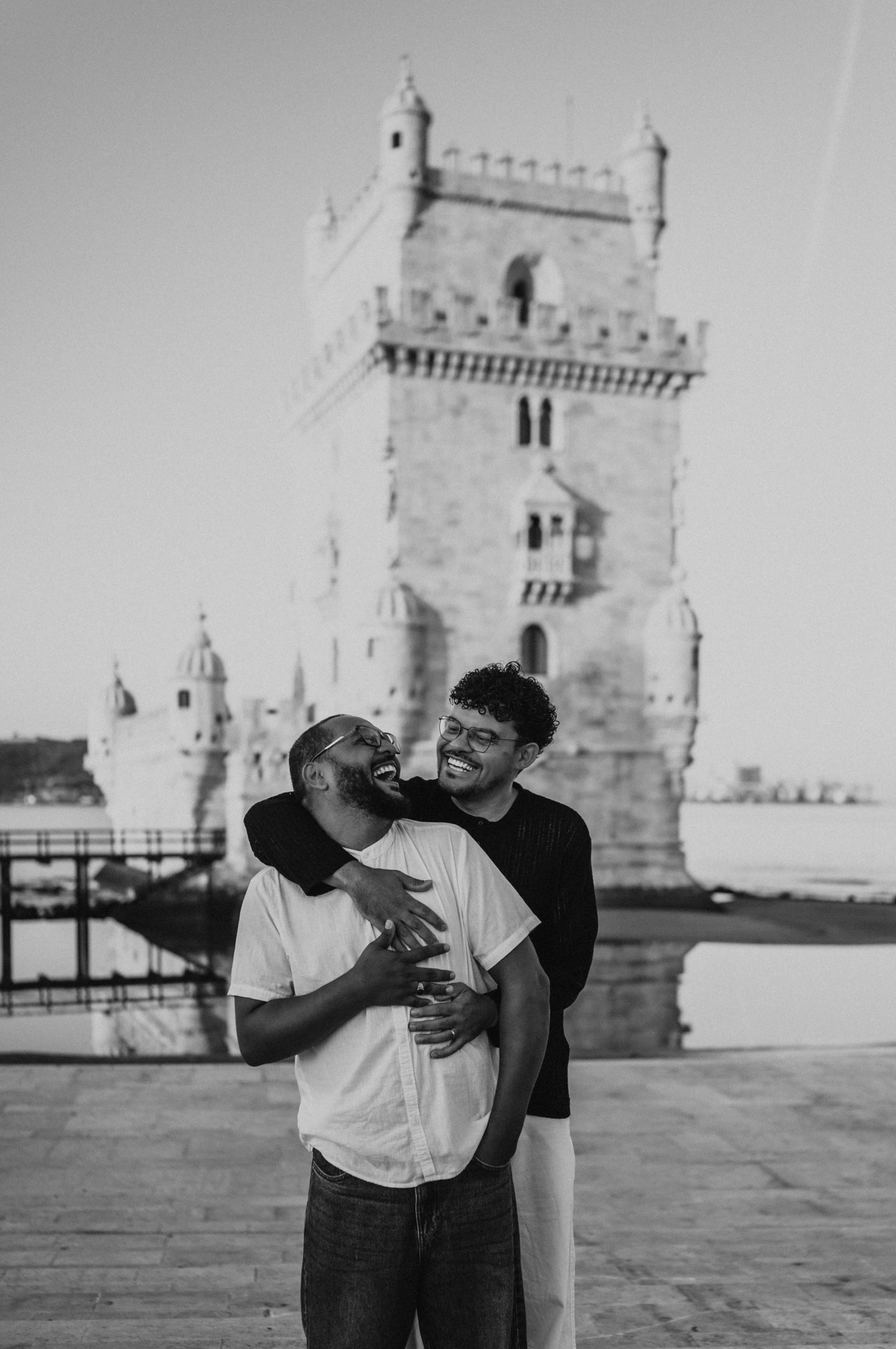 A black-and-white portrait of the couple smiling at each other in an embrace by Belém Tower during their couple photoshoot in Lisbon.