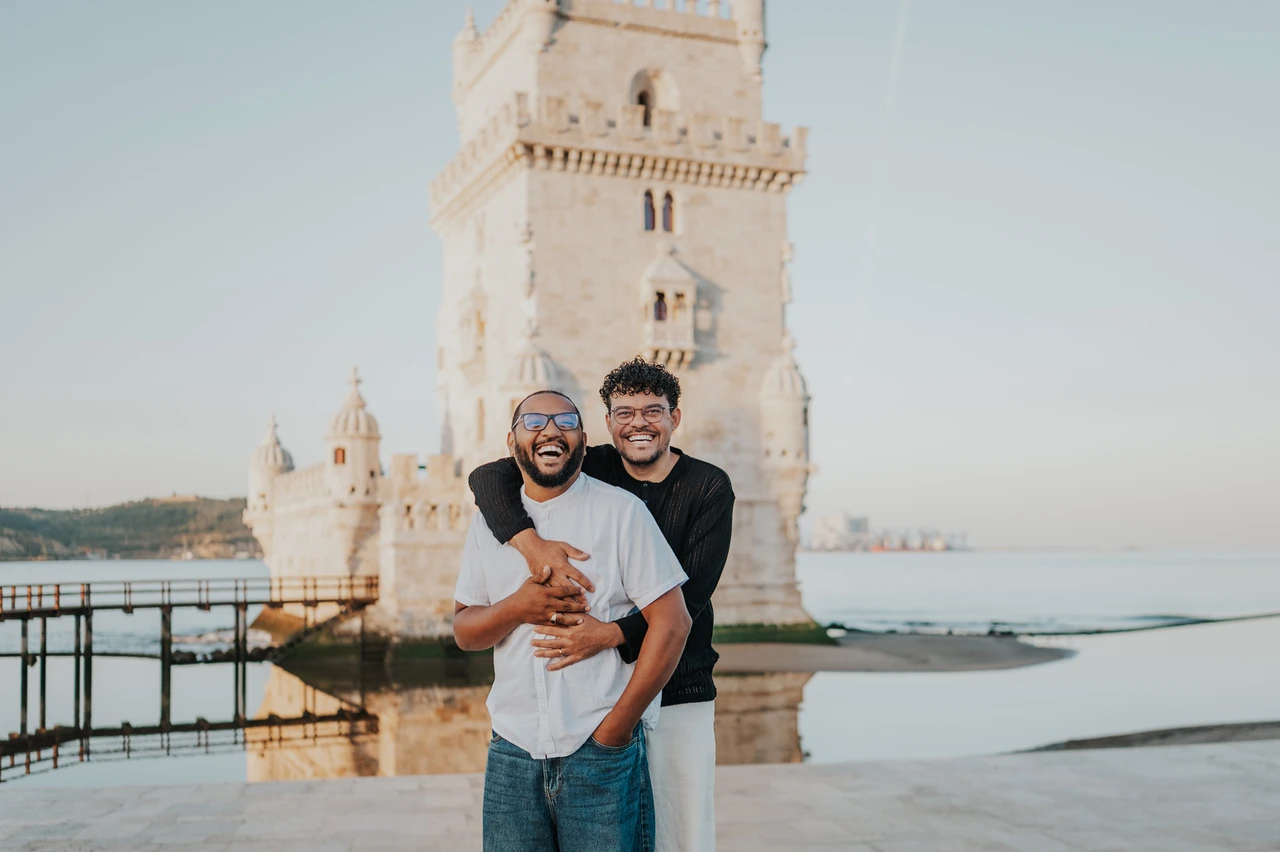 A smiling landscape portrait at the tower | Lisbon couple photoshoot A landscape portrait of the couple smiling together in an embrace with Belém Tower behind them during their couple photoshoot in Lisbon.