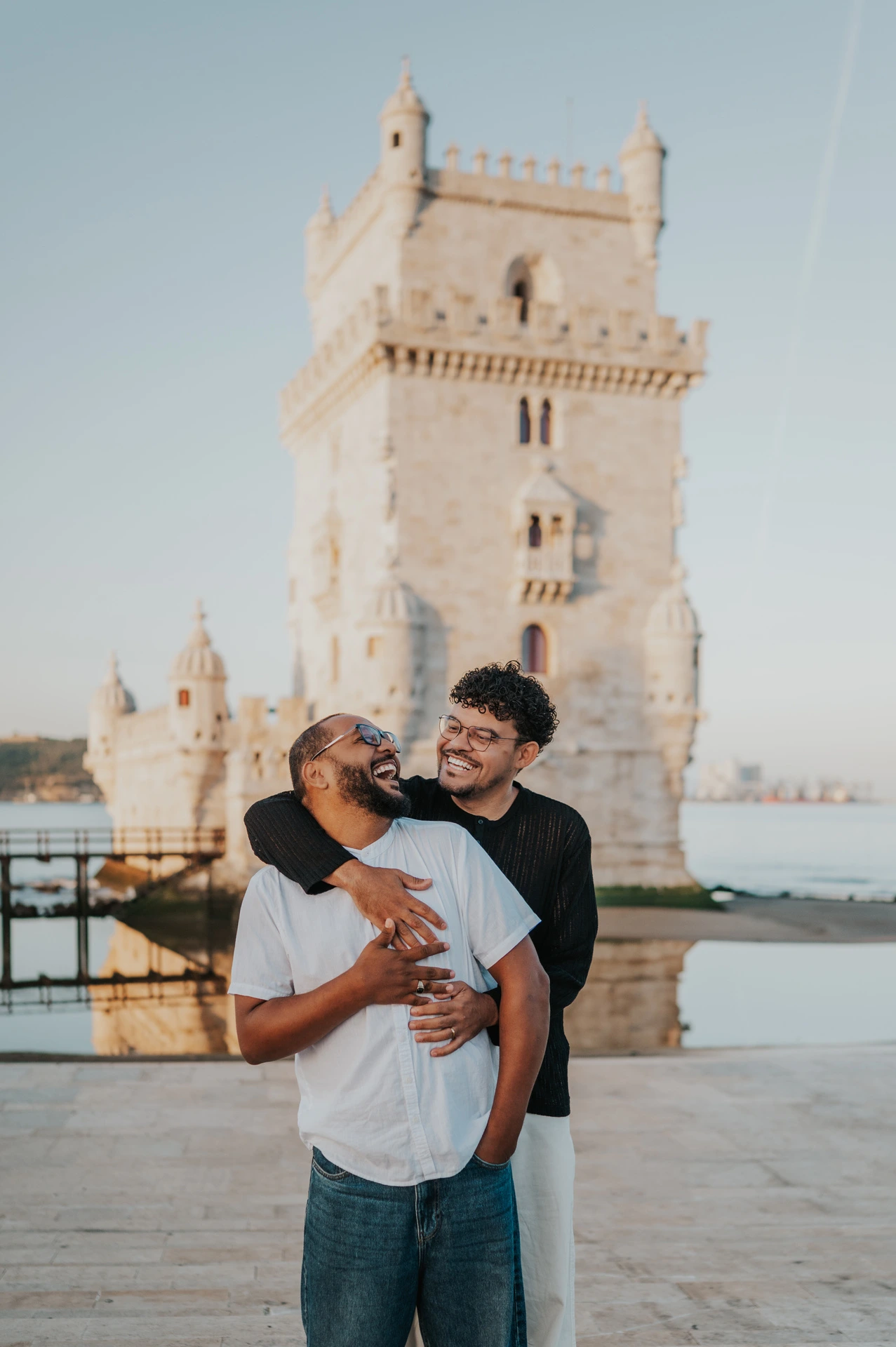The couple looking at each other and smiling in an embrace with Belém Tower behind them during their couple photoshoot in Lisbon.