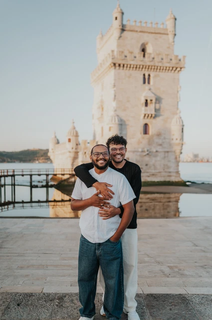 Smiling together in an embrace | Lisbon couple photoshoot The couple smiling towards the camera with one partner embracing the other from behind by Belém Tower during their couple photoshoot in Lisbon.