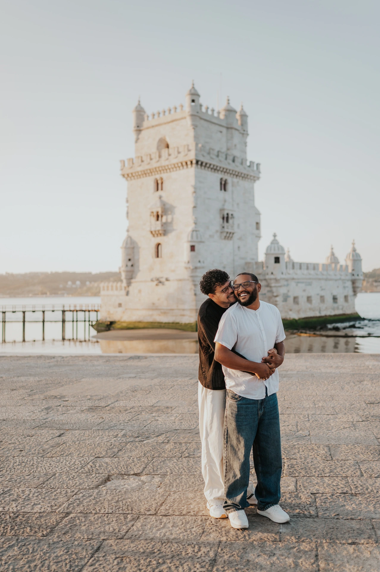 A full-length portrait of the couple holding each other in front of Belém Tower during their couple photoshoot in Lisbon.