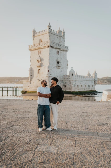 A cheek kiss on the waterfront | Lisbon couple photoshoot One partner kissing the other on the cheek while holding him close in front of Belém Tower during their couple photoshoot in Lisbon.
