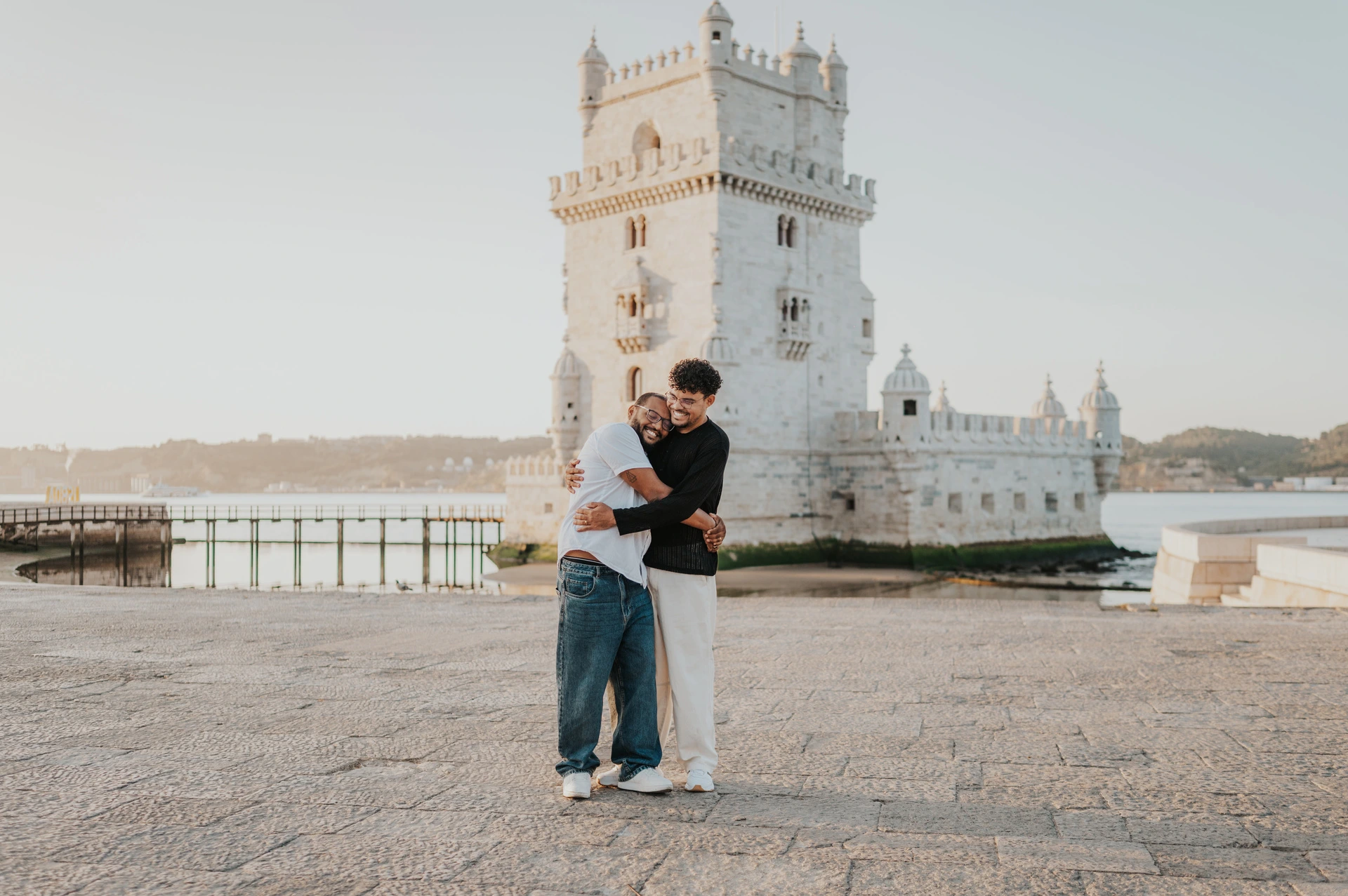A smiling embrace on the waterfront | Lisbon couple photoshoot A landscape portrait of the couple holding each other close and smiling beside Belém Tower during their couple photoshoot in Lisbon.