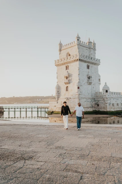 A wide waterfront walk | Lisbon couple photoshoot A wide portrait of the couple walking together across the paving beside Belém Tower during their couple photoshoot in Lisbon.